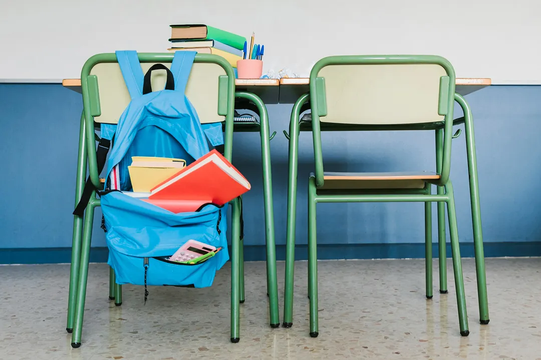 Two school chairs and desks with a blue satchel on the back, full of books.